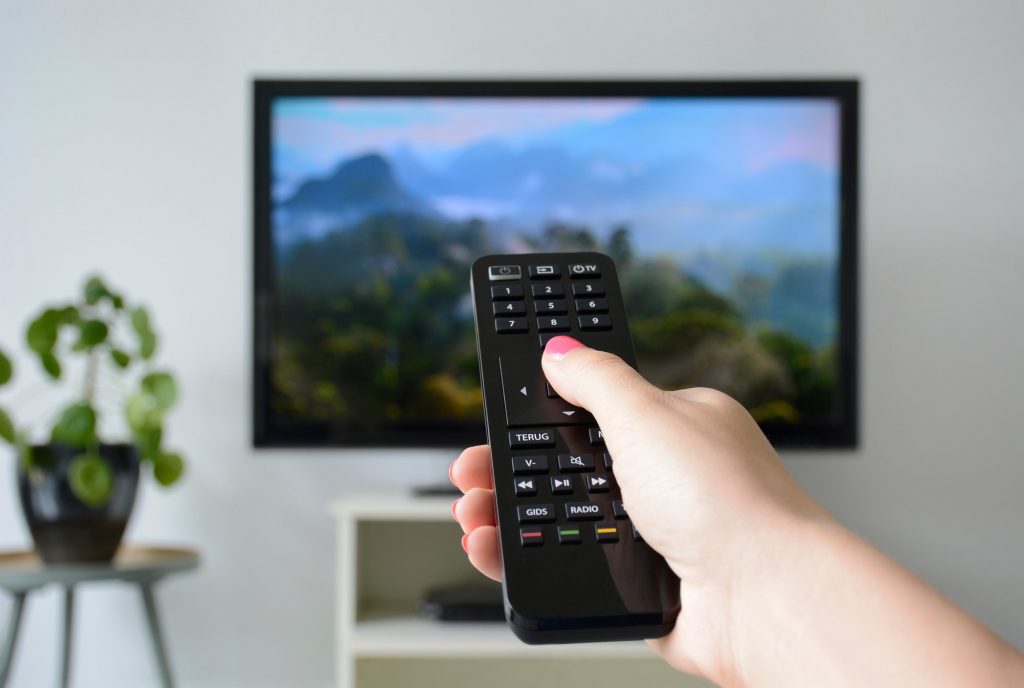 Watching tv. A young woman’s hand holding the tv remote control with a television in the background